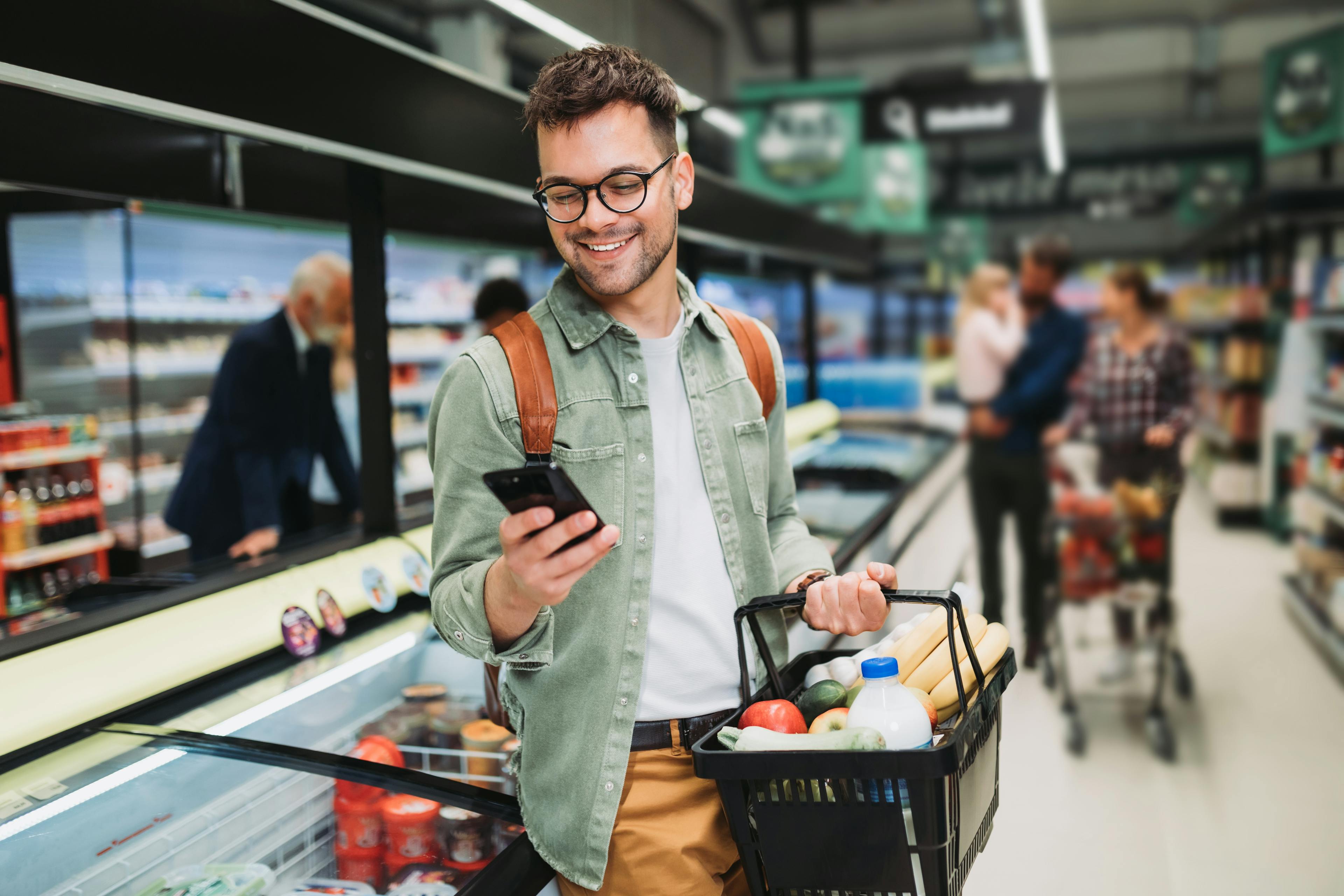 young man in grocery store holding a shopping basket and looking at his phone